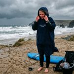 Beginner surfer wrapping up in a hooded surf poncho on a cold, windy beach after a session