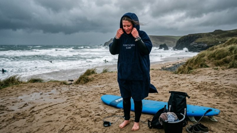 Beginner surfer wrapping up in a hooded surf poncho on a cold, windy beach after a session