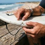 Surfer inspecting a small fiberglass ding on a surfboard rail outdoors.